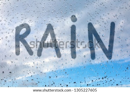 The word rain is written by a finger on the misted glass. Window glass with raindrops against a blue sky and sun. Imitation of children's inscriptions.