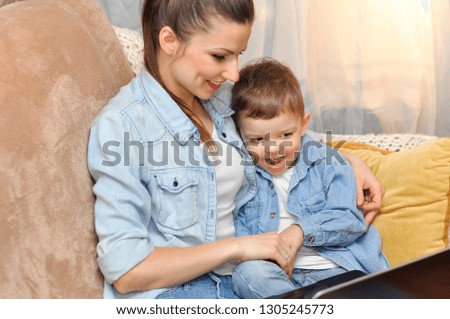 A young mother with her baby son are lying on the couch together in the same clothes and look at the laptop.