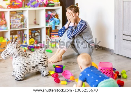 Tired of everyday household mother sitting on floor with hands on face. Kid playing in messy room. Scaterred toys and disorder. Happy parenting Royalty-Free Stock Photo #1305255670