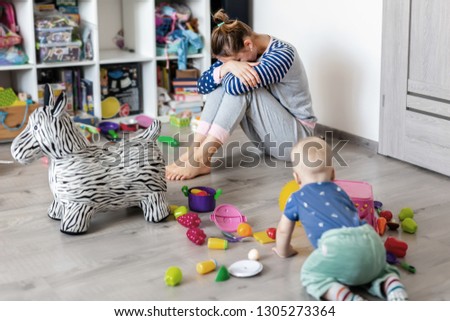Tired of everyday household mother sitting on floor with hands on face. Kid playing in messy room. Scaterred toys and disorder. Happy parenting Royalty-Free Stock Photo #1305273364
