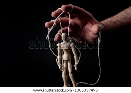 Employer manipulating the employee, emotional manipulation and obey the master concept with ominous hand pulling the strings on a marionette with moody contrast on black background Royalty-Free Stock Photo #1305339493