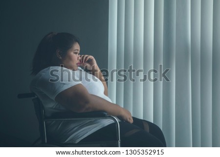 Picture of overweight woman looks sad while sitting in the wheelchair by the window