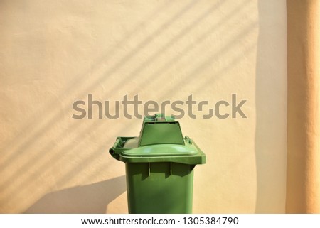 The large green trash can be placed in the middle of the picture, with a cream colored cement wall with cement pillars with beautiful light and shadow