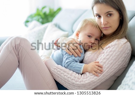 Young mother, holding her sick toddler boy, hugging him at home, sunny living room
