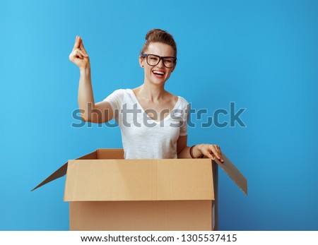 happy modern woman in white t-shirt with fingers snapping in a cardboard box isolated on blue background Royalty-Free Stock Photo #1305537415