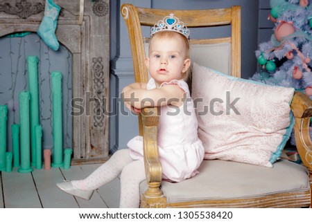 Portrait of cute little european blond princess girl with a crown  in a beautiful dress sitting on a vintage chair in a studio decorated in the Christmas theme with toys, tree and fireplace