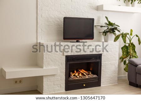 Room in a modern style. There is a TV on the white wall, white rack with a flower in vase, TV remote, decorations in a form black fireplace