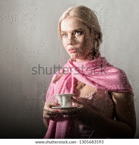 young blonde woman behind glass with water drops. beautiful girl drinks coffee or tea
