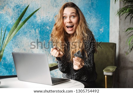 Picture of beautiful excited young pretty woman sitting in cafe indoors using laptop computer holding credit card.