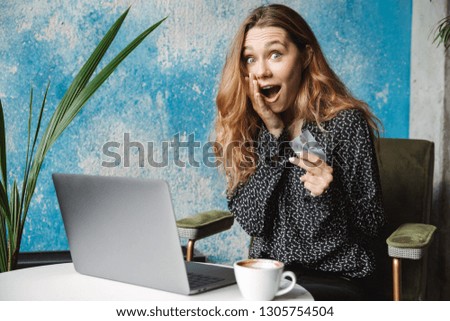Photo of excited emotional beautiful young pretty woman sitting in cafe indoors using laptop computer drinking coffee holding credit card.