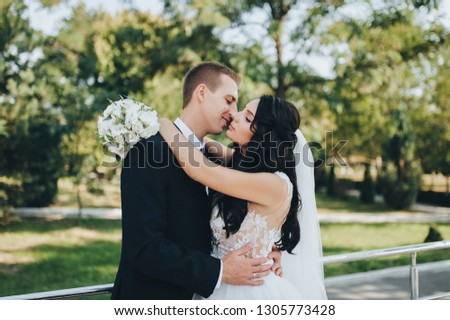 Beautiful brunette bride in a white dress with a bouquet and stylish groom in a black suit cuddling in the garden with greenery. Portrait of lovers and smiling newlyweds. Wedding photography.