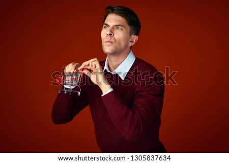 A puzzled man in a red sweater holds an iron cart in his hands on a red background
