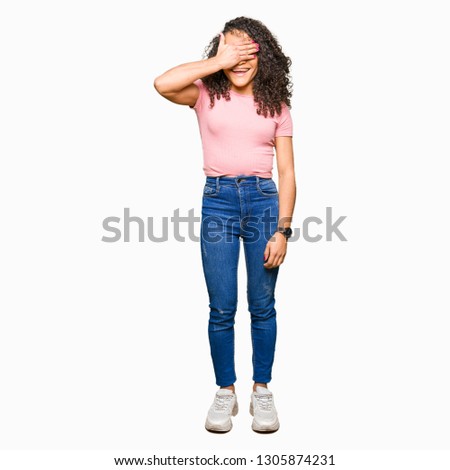 Young beautiful woman with curly hair wearing pink t-shirt smiling and laughing with hand on face covering eyes for surprise. Blind concept.