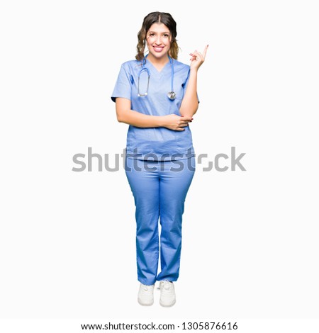 Young adult doctor woman wearing medical uniform with a big smile on face, pointing with hand and finger to the side looking at the camera.