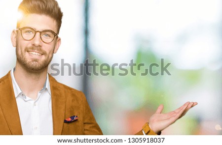 Young handsome business man wearing glasses smiling cheerful presenting and pointing with palm of hand looking at the camera.