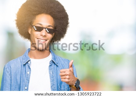 Young african american man with afro hair wearing thug life glasses doing happy thumbs up gesture with hand. Approving expression looking at the camera with showing success.