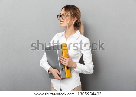 Photo of caucasian businesswoman wearing glasses holding paper folders in the office isolated over gray background