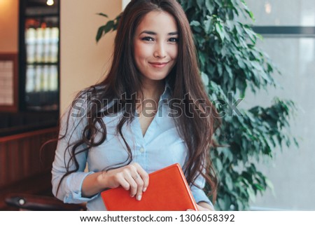 Portrait of beautiful charming brunette long hair smiling asian girl with orange leather diary near the window at cafe