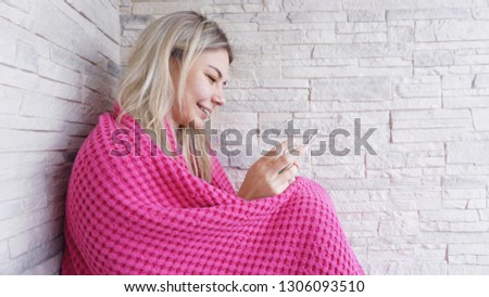 Pretty girl sitting on the window sill with smartphone in hands. She has long blonde hair, smile and looking at her phone. Pink plaid