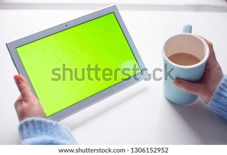 Woman hand hold white tablet with green screen and coffee cup in office on white table. Close up.