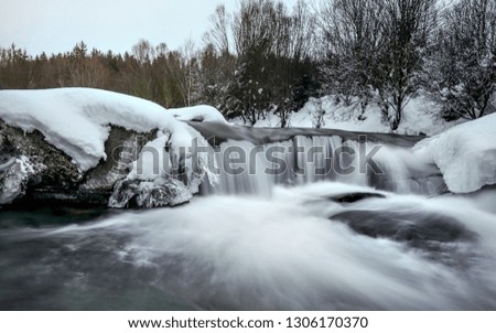 Small waterfall on river in winter, long exposure makes water milky smooth, snow and ice around, morning sun setting up distant sky.