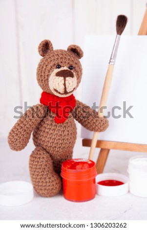Knitted brown bear with brush and paint near the easel. Handwork, creativity. Amigurumi. Postcard