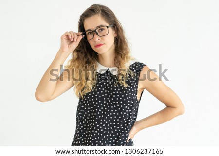 Serious woman adjusting glasses and keeping hand on hip. Beautiful young lady standing and posing at camera. Woman portrait concept. Isolated front view on white background.
