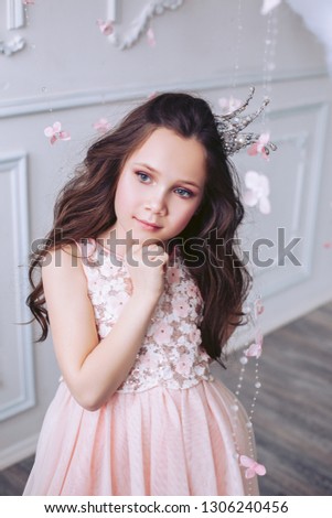 Cute little girl with a dreamy facial expression posing against a white wall in a beautiful pink dress and a crown on her head.