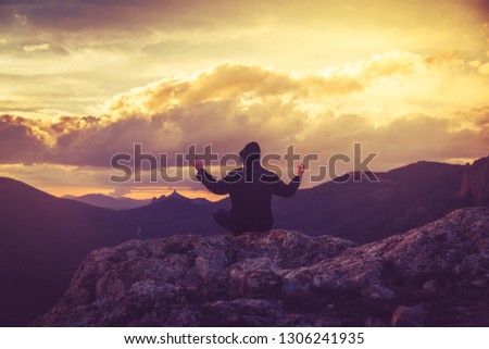 A young man is meditating in the lotus position on top of a mountain against the backdrop of a sunset, view from the back