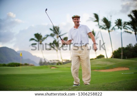 Male golfer smoking a cigar holds his golf club aloft as he poses for a portrait on a golf course.