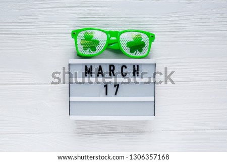 Creative festive St Patricks background with white light box calendar 17 march and fun green glasses with shamrock on white wooden table. Flat lay composition, copy space.