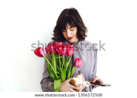 Beautiful happy girl with bouquet of tulips and gift box on white background indoors, space for text. Stylish young woman holding phone and flowers.  International womens day. Hello spring