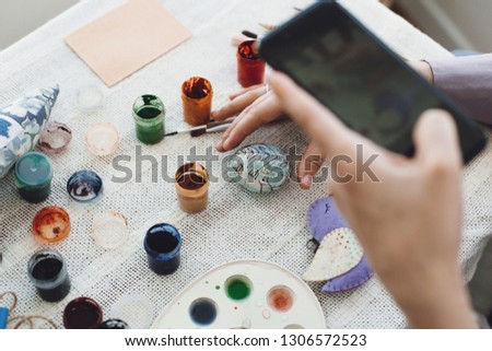 Hands holding phone and taking photo of stylish easter flat lay of painting egg on rustic table with paint,brushes, bird toy. Easter workshop.Decorating easter egg, holiday preparations.