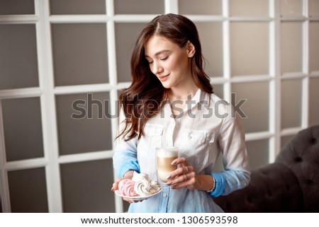 Beautiful young woman with dark hair dressed in a white shirt is standing in a cafe indoors with a glass cup of latte on soy milk.