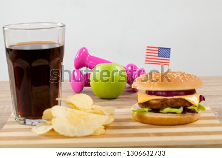 Close up picture of tasty double cheeseburger, potato chips and a glass of soda - fast food. Dumbbells and green apple in the background