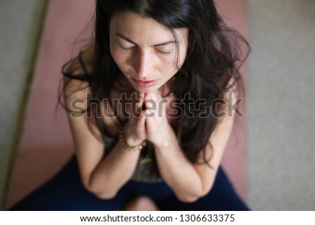Gorgeous young Europian female on dark background, holding hands in namaste or prayer,  keeping eyes closed while practising yoga and meditating at home alone, having calm look on her face. Top view
