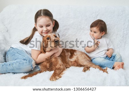 Two baby girls, sisters play on white sofa with red dog.