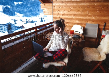 Smart and beautiful girl sitting on the balcony of the hotel in winter. She is working in internet, holding the laptop on her knees. Wearing red pajama pants and creamy cweater. Amazing mountains view