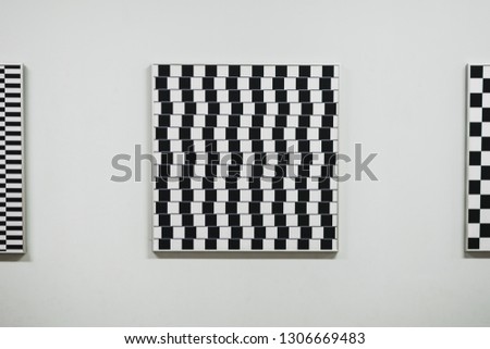 Picture with black and white square pattern hanging on the white wall. Left and right are two part of similar pictures. Museum interior. 
