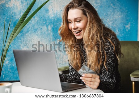Picture of beautiful young pretty woman sitting in cafe indoors using laptop computer holding credit card.