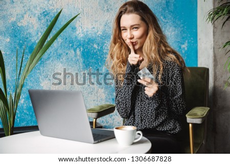 Picture of beautiful young pretty woman sitting in cafe indoors using laptop computer drinking coffee showing silence gesture holding credit card.