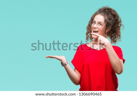 Beautiful brunette curly hair young girl wearing glasses over isolated background amazed and smiling to the camera while presenting with hand and pointing with finger.