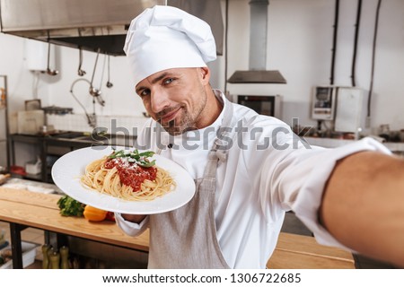Image of handsome man chief in apron taking selfie while standing at kitchen in restaurant