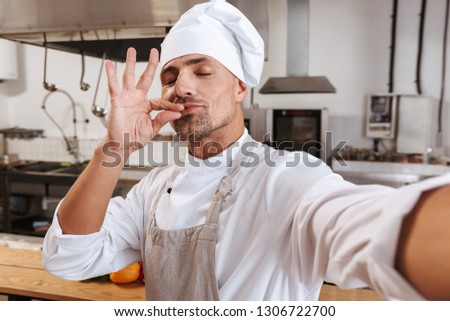 Photo of caucasian male chief in apron taking selfie while standing at kitchen in restaurant