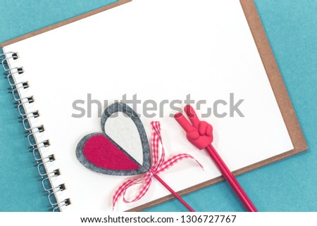 A red pencil with V sign, red crafted heart over a blank spiral notepad or scetch book on blue background. Flat lay with copy space