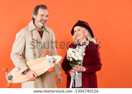 cheerful retired woman with flowers standing near husband with skateboard isolated on orange