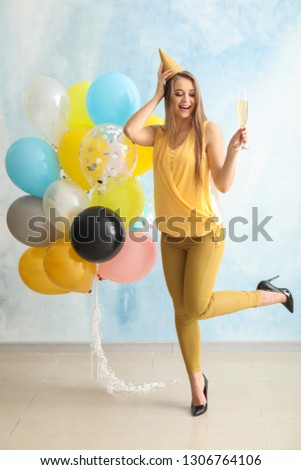 Beautiful young woman with balloons, party hat and glass of champagne near light wall