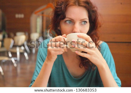 Nice redhead lady shot relaxed in the cafeteria while drinking from a cup of tea. She looks very meditative and pensive while looking away the camera. There's a blurry cafeteria  interior behind her