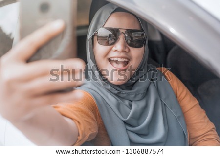 Portrait of Asian muslim lady smiling while taking selfie photo picture with smart phone in her car 