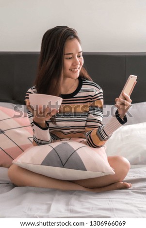 Beautiful young Asian girl on the bed holding mobile phone eating snacks